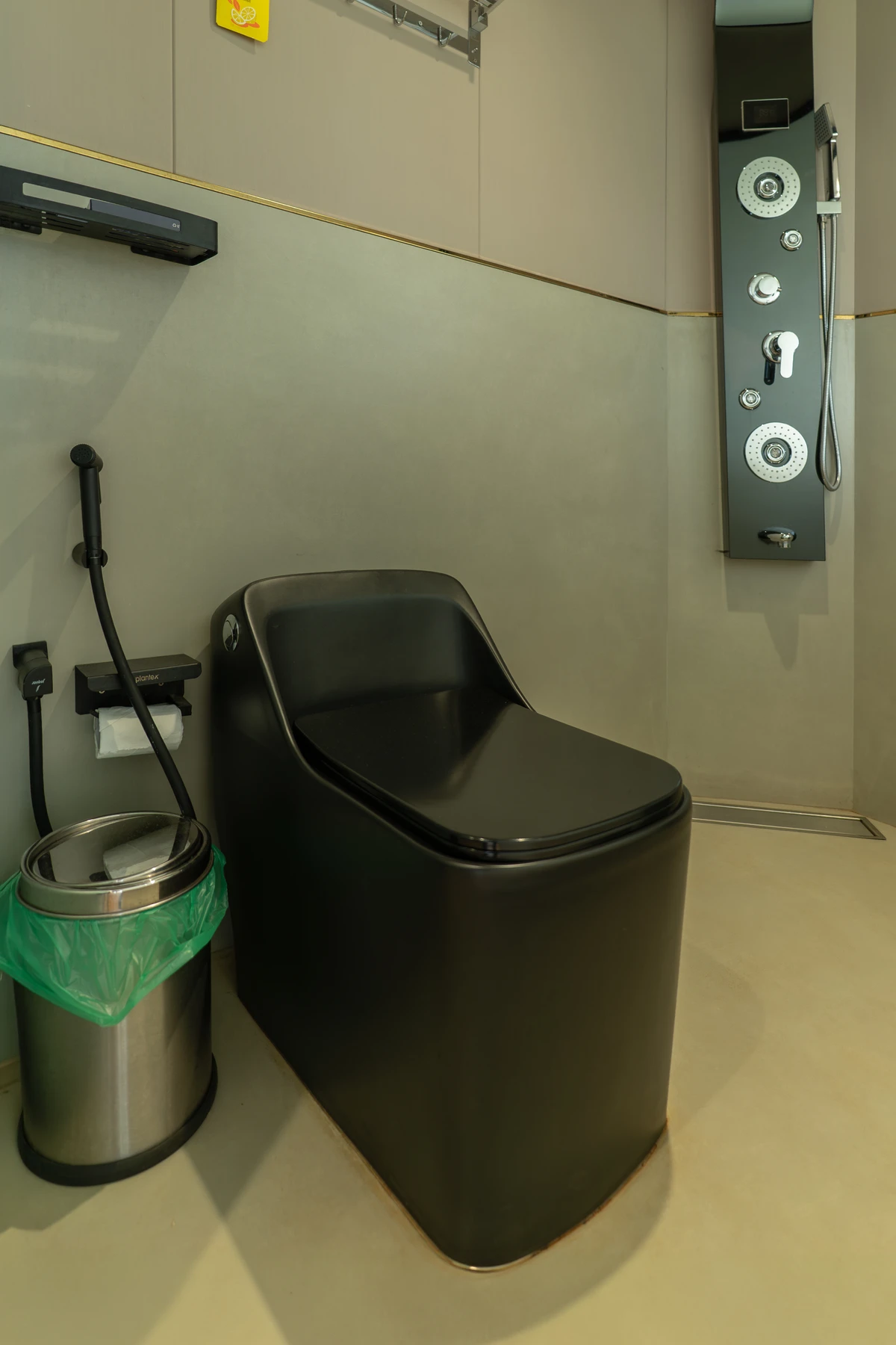 A modern bathroom stall featuring a matte black toilet, a stainless steel trash can, and a black multi-function shower panel against minimalist beige walls.